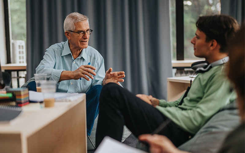 A mature man with grey hair and glasses sits next to a desk speaking with two younger people, one a male with a green shirt and the other blurred in the foreground